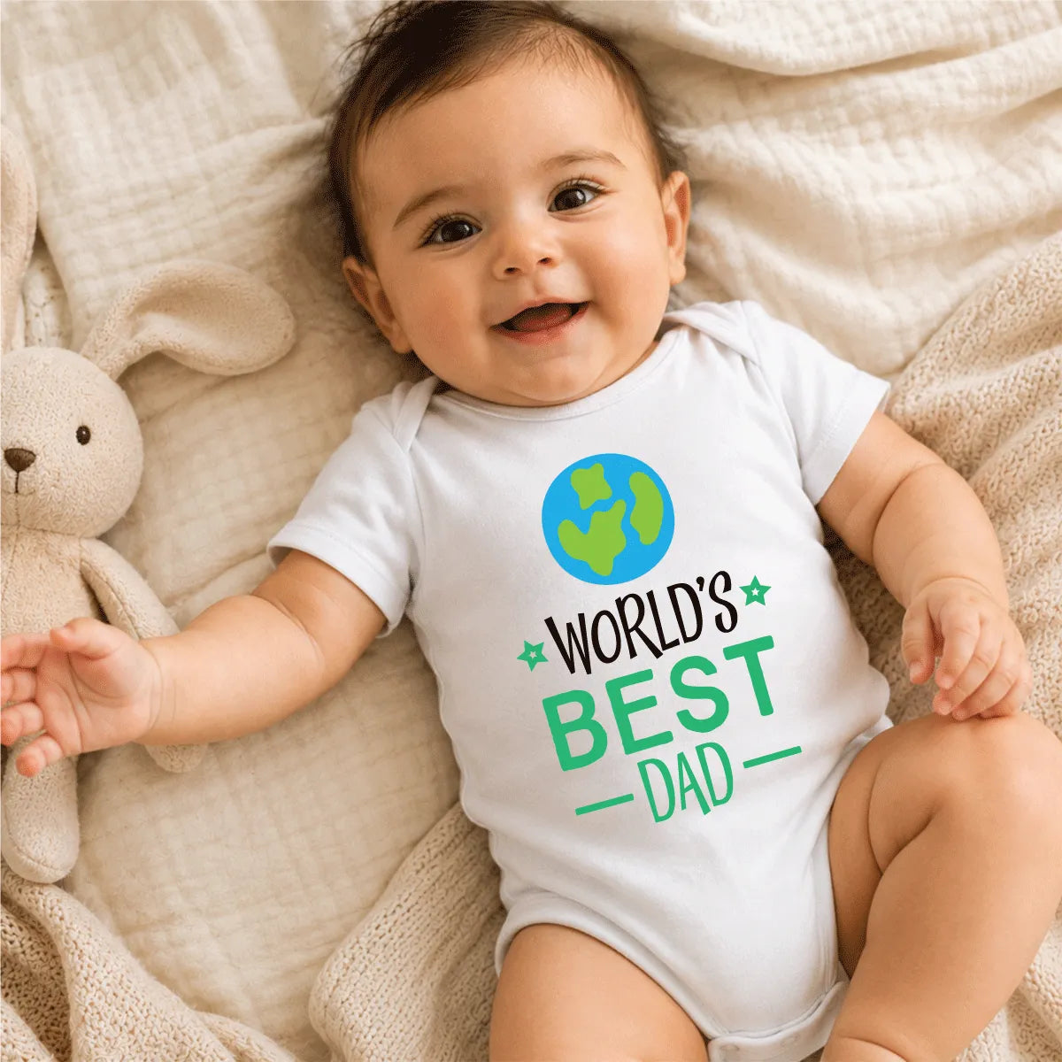Baby wearing a 'World's Best Dad' onesie lying on a soft surface with a teddy bear.